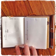 a person holding an open notebook on top of a wooden table next to a pen