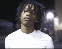 a man with dreadlocks standing in front of a building at night, wearing a white t - shirt