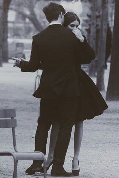 a man and woman standing next to each other near a bench in a park with trees