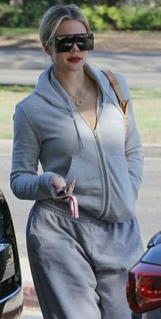 a woman in grey sweatsuit and sunglasses walking down the street with her cell phone