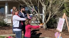 a group of people standing in front of a house holding up balloons and pointing them at an easel