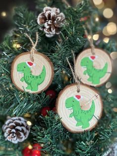 three wooden ornaments with green dinosaurs on them are hanging from a christmas tree in front of some pine cones