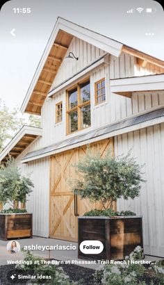 a large white barn with wooden doors and plants in the front yard, on a sunny day