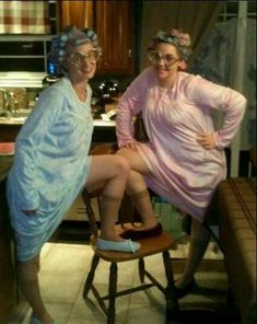 two women in costumes sitting on stools with their legs crossed and one wearing glasses