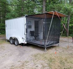 an enclosed trailer sits in the middle of a wooded area with chairs and trees around it