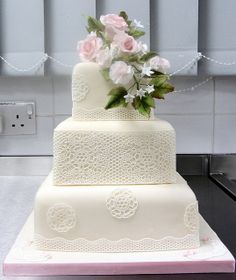 a three tiered wedding cake with pink roses on top and white lace around the edges