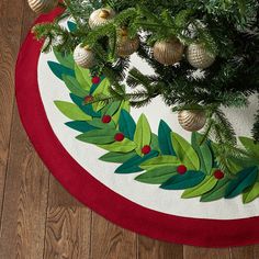 a christmas tree skirt with green leaves and red berries on the bottom is shown in front of a wooden floor