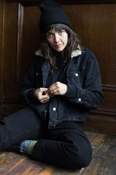 a woman sitting on the floor with her hands in her pockets and wearing a black hat