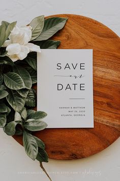 a save the date card sitting on top of a wooden plate with greenery around it