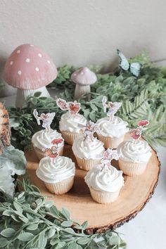 cupcakes with white frosting sitting on a wooden plate in front of mushrooms and greenery