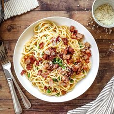 a white bowl filled with pasta and bacon on top of a wooden table next to silverware