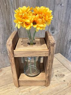 some yellow flowers are in a glass vase on a wooden stand with wood planks