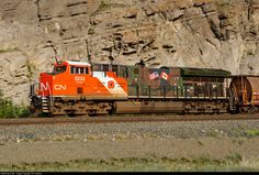 an orange and white train traveling past a rocky mountain side covered in graffiti on the side of it