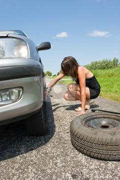 a woman kneeling down next to a car on the side of the road with a tire in front of her