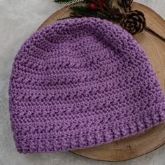 a purple crocheted hat sitting on top of a wooden plate next to a pine cone