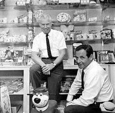 two men sitting in front of a store with stuffed animals on the floor and shelves