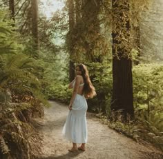 a woman in a white dress is walking down a path through the woods with trees