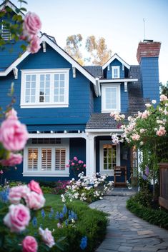 a blue house with white windows and lots of pink flowers in the front yard on a sunny day