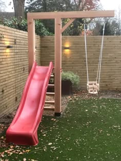 a red slide is in the grass next to a wooden swing set and brick wall