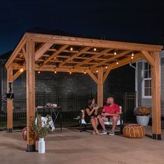 two people sitting on a bench under a wooden pergolan with lights strung over them