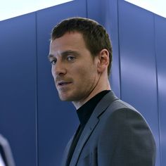 a man standing in front of a blue wall wearing a gray suit and black turtle neck sweater