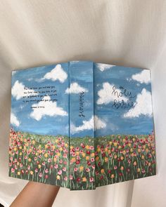 a person holding up a book with an image of flowers and clouds painted on it