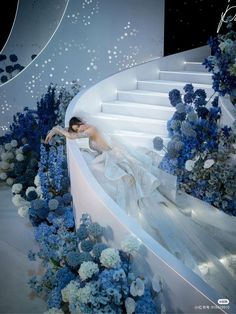 a woman in a white dress is laying down on a staircase surrounded by blue and white flowers