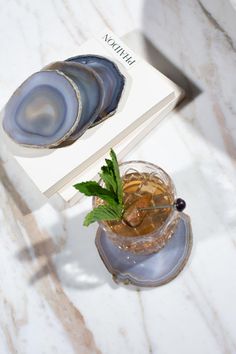 a book and some drinks on a white marble counter top with a leafy green garnish