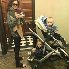 a woman standing next to a baby in a stroller and holding a cell phone