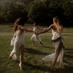 two women in white dresses holding hands and dancing on grass with trees in the background