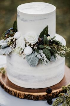 a white wedding cake with flowers and greenery on top is sitting on a wooden slice