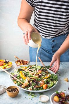 a woman pouring dressing onto a salad in a white bowl on top of a table