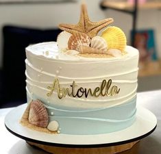 there is a white cake with shells on the top and name written in gold lettering