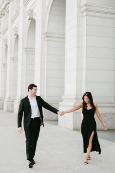 an engaged couple holding hands and walking down the street in front of white pillars with arches