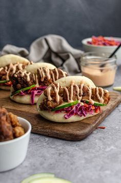 three tasty looking sandwiches on a cutting board next to bowls of sauce and condiments