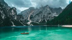 two people in a small boat on a lake surrounded by mountains and pine trees, under cloudy skies