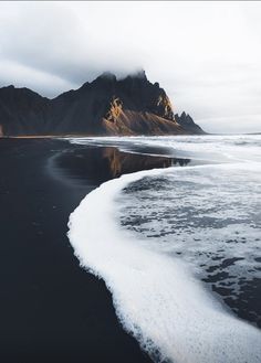 the black sand beach has white foam on it and mountains in the backgroud