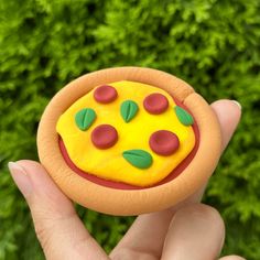 a hand holding a small toy pizza with toppings on it's face in front of some bushes