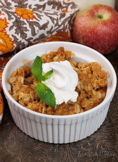 an apple crisp with whipped cream in a white bowl next to two apples on a table