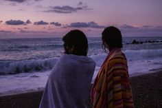 two people sitting on the beach watching the waves come in to shore as the sun sets