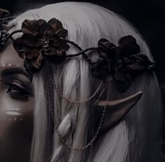 a woman with long white hair wearing horns and flowers on her head, in front of a mannequin's face