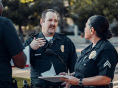 two police officers talking to each other in front of a microphone and another officer holding a clipboard