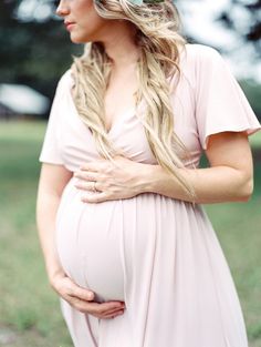 a pregnant woman wearing a flower in her hair standing on the grass with trees behind her