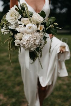 a bride holding a bouquet of white flowers and greenery in her hand while standing on the grass