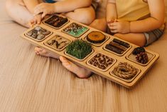 two children sitting on a bed holding a wooden tray with different items in it,