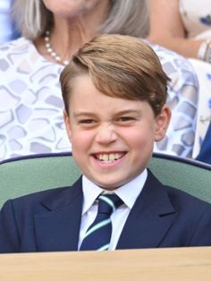 a young boy wearing a suit and tie sitting in a chair with an older woman behind him