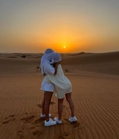 two people standing in the desert with their arms around each other as the sun sets