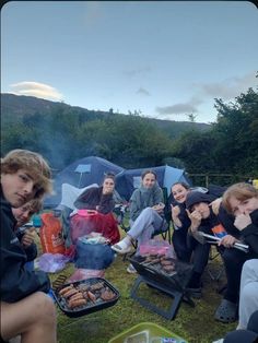 a group of people sitting around a campfire with food on the grill and tents in the background