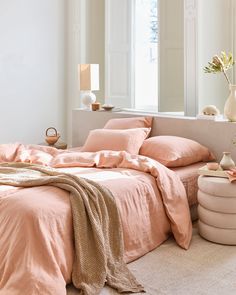 a bed with pink sheets and pillows in a white room next to a large window