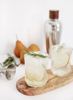 two glasses filled with ice sitting on top of a cutting board next to pears